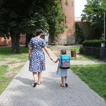 Eine erwachsene Person und ein Kind mit Schulmappe sind von hinten zu sehen und laufen im Park.