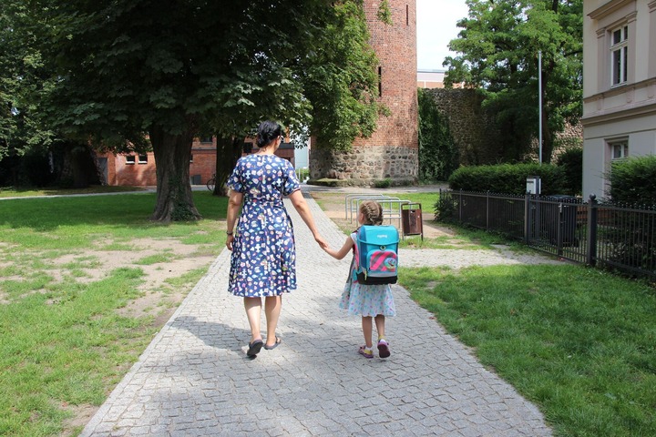 Eine erwachsene Person und ein Kind mit Schulmappe sind von hinten zu sehen und laufen im Park.