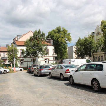 Unbefestigter Schotterparkplatz, beidseitig vollständig mit Autos belegt, dahinter Wohnhäuser mit Altbaufassaden und Bäume unter bewölktem Himmel.