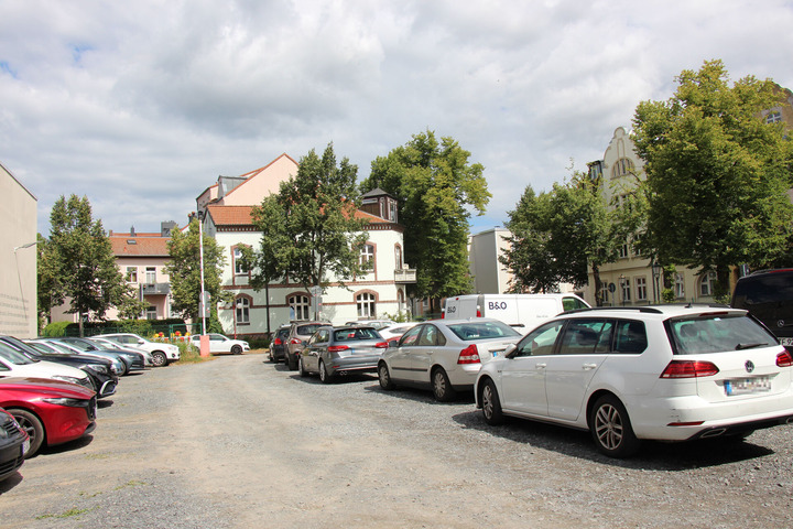 Unbefestigter Schotterparkplatz, beidseitig vollständig mit Autos belegt, dahinter Wohnhäuser mit Altbaufassaden und Bäume unter bewölktem Himmel.