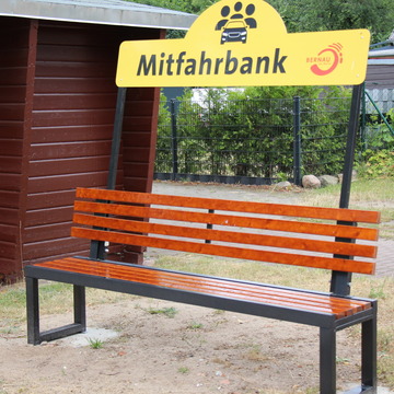Eine Bank mit einem Schild auf dem "Mitfahrbank" steht, steht direkt neben einem Bushaltestellenhäuschen. 