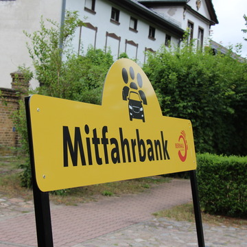 Nahaufnahme des Schildes über der Mitfahrbank. Darauf steht das Wort "Mitfahrbank"