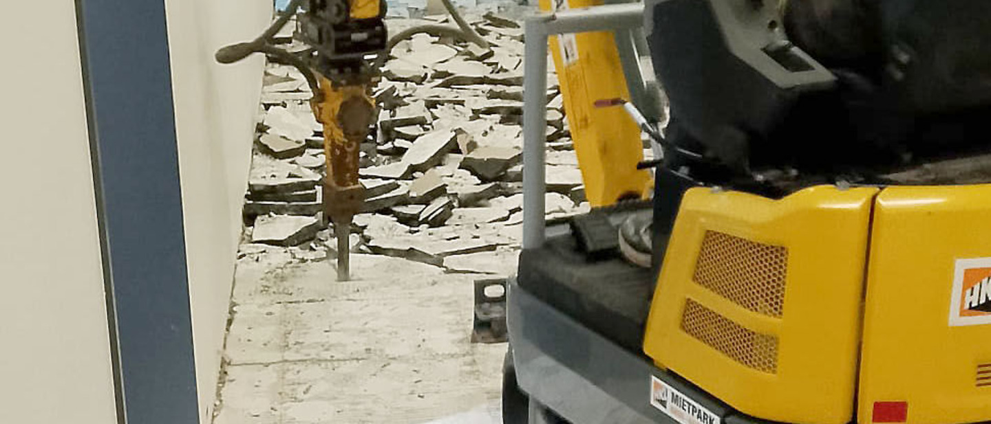 Eine Baumaschine wird von einem mann bedient. Der Boden im Schulflur wird aufgestemmt.