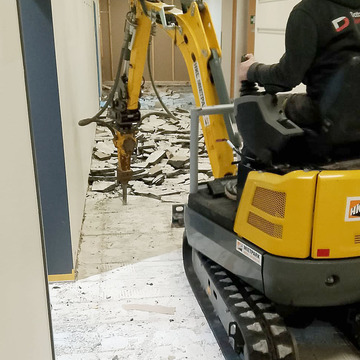 Eine Baumaschine wird von einem mann bedient. Der Boden im Schulflur wird aufgestemmt.