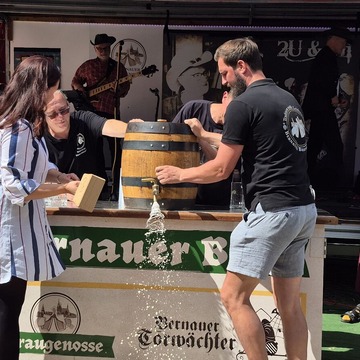 Vier Personen stehen um ein Bierfass herum. Eine von ihnen sticht das Bierfass an. 