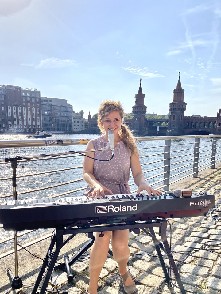 Eine Sängerin steht an einem Keyboard und singt. Im Hintergrund ist Wasser zu sehen.