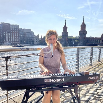 Eine Sängerin steht an einem Keyboard und singt. Im Hintergrund ist Wasser zu sehen.