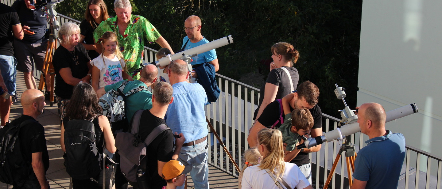 Menschen stehen auf der Dachterrasse und schauen durch Teleskope in Richtung Sonne.
