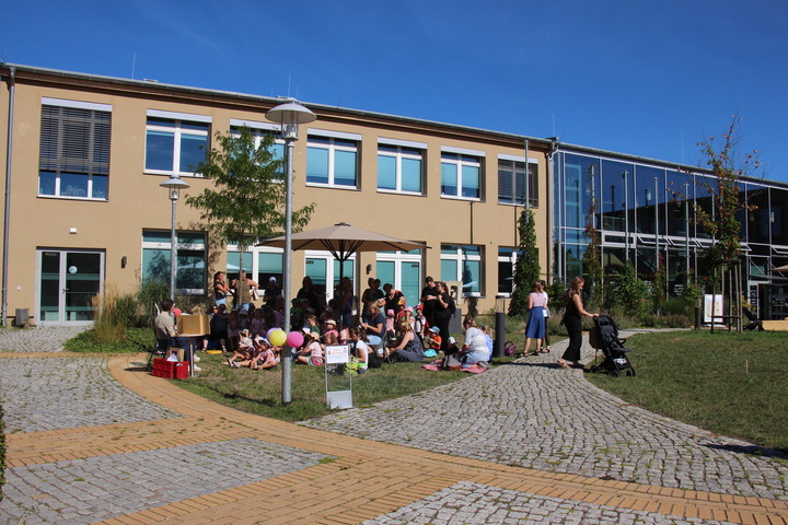 Unter einem Sonnenschirm sitzen mehrere Kinder und lauschen einer Geschichte, die vorgelesen wird. 