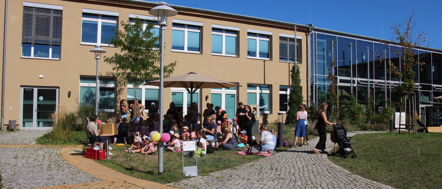 Unter einem Sonnenschirm sitzen mehrere Kinder und lauschen einer Geschichte, die vorgelesen wird. 