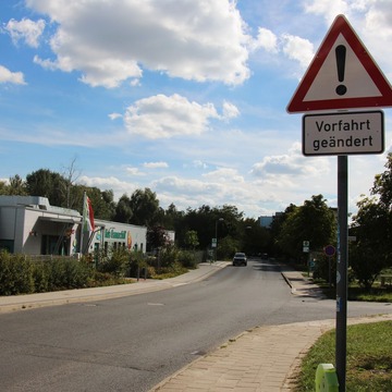 Eine Straße mit einem Verkehrsschild. Darauf ist ein Ausrufezeichen und die Info "Vorfahrt geändert" zu sehen. IM Hintergrund ist die Kreuzung und eine Kita zu sehen. 