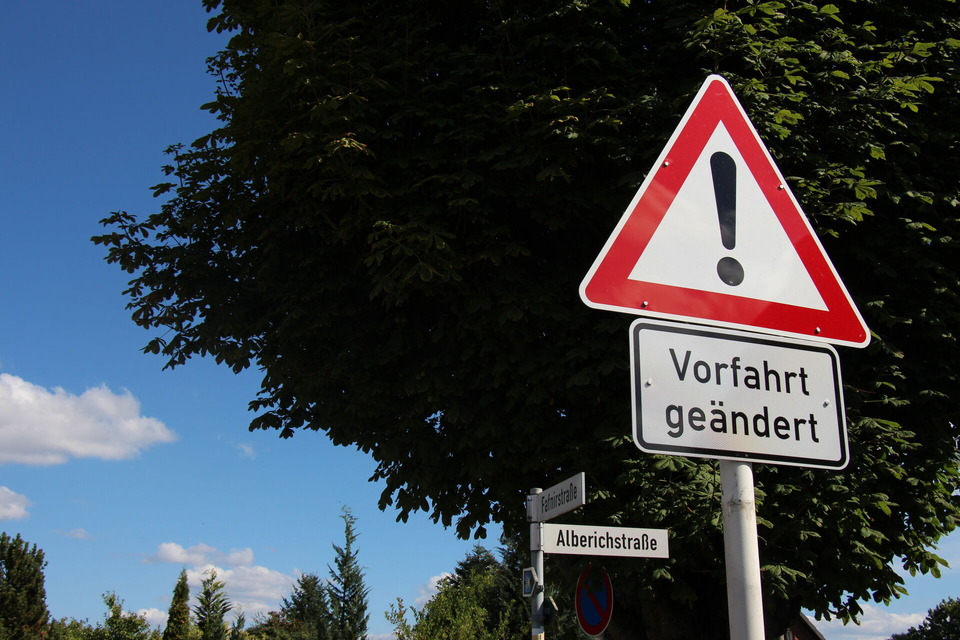 Nahaufnahme des Verkehrsschilder mit AUsrufezeichen und "Vorfahrt geändert", dahinter ist das Straßennamenschild der "Alberichstraße" zu sehen.