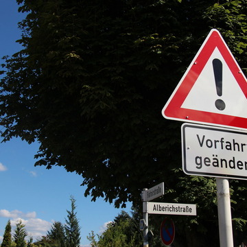Nahaufnahme des Verkehrsschilder mit AUsrufezeichen und "Vorfahrt geändert", dahinter ist das Straßennamenschild der "Alberichstraße" zu sehen.