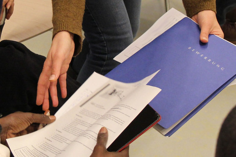Nur die Hände von zwei Personen sind sichtbar. Eine Mappe mit der Aufschrift "Bewerbung" wird überreicht. 