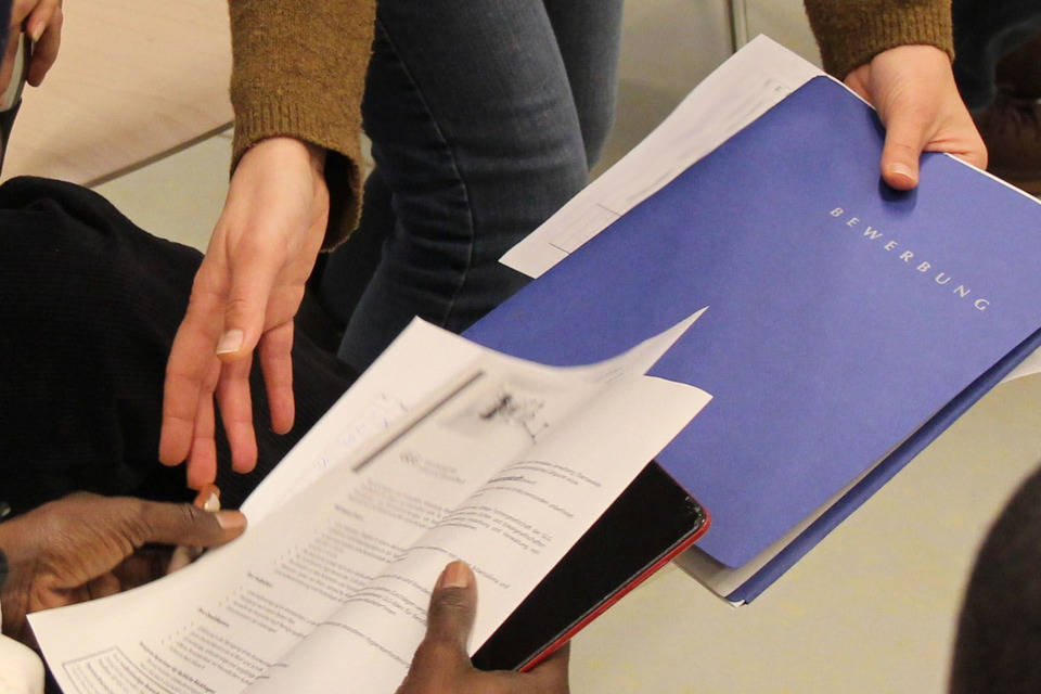 Nur die Hände von zwei Personen sind sichtbar. Eine Mappe mit der Aufschrift "Bewerbung" wird überreicht. 