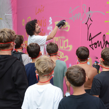 Workshopleiter zeigt wie gesprayt wird, Kinder und Jugendliche schauen zu.