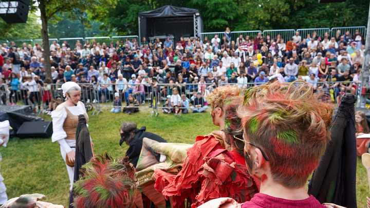 Man sieht viele interessierte Zuschauende auf Tribünen, die sich ein Theaterstück im Bernauer Stadtpark schauen.