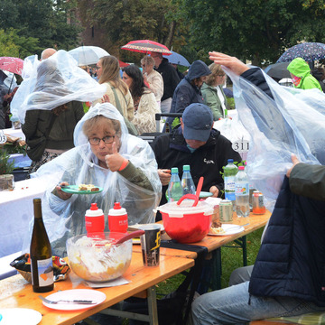 An Bierbänken sitzen mehrere Menschen in Regenponchos und speisen. Eine Frau hält den Daumen nach oben. 