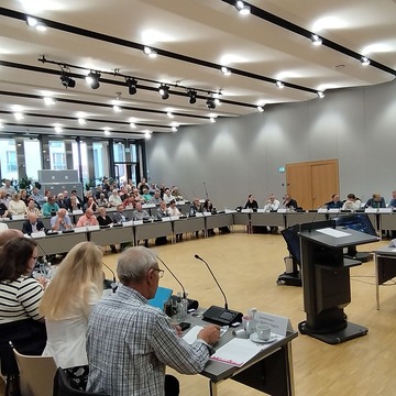 Das Bild zeigt die Stadtverordnetenversammlung, welche sich in einem großen Saal berät 
