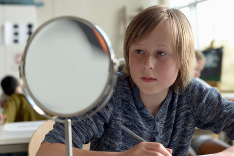 Das Bild zeigt einen Jungen, welcher an einem Schultisch sitzt und in einen kleinen Spiegel schaut