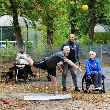 Mann beim Kugelstoßen, im Hintergrung der Prüfer im Rollstuhl.