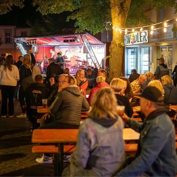 Mehrere Bierbänke und Tische stehen vor einer Bühne. Lichterketten über den Köpfen mehrerer Gäste.