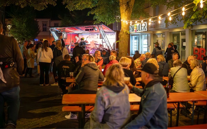 Mehrere Bierbänke und Tische stehen vor einer Bühne. Lichterketten über den Köpfen mehrerer Gäste.