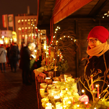 Auf einem Weihnachtsmarkt steht ein Stand an dem Gläser und Gefäße für erzen angeboten werden. Die Händlerin am Stand lächelt in die Kamera